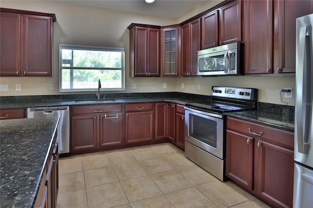 kitchen with stainless steel appliances, a sink, dark stone counters, glass insert cabinets, and light tile patterned floors