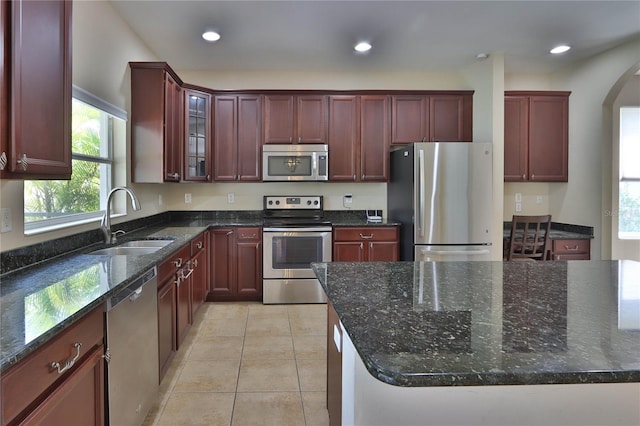 kitchen with stainless steel appliances, dark stone countertops, reddish brown cabinets, a sink, and arched walkways