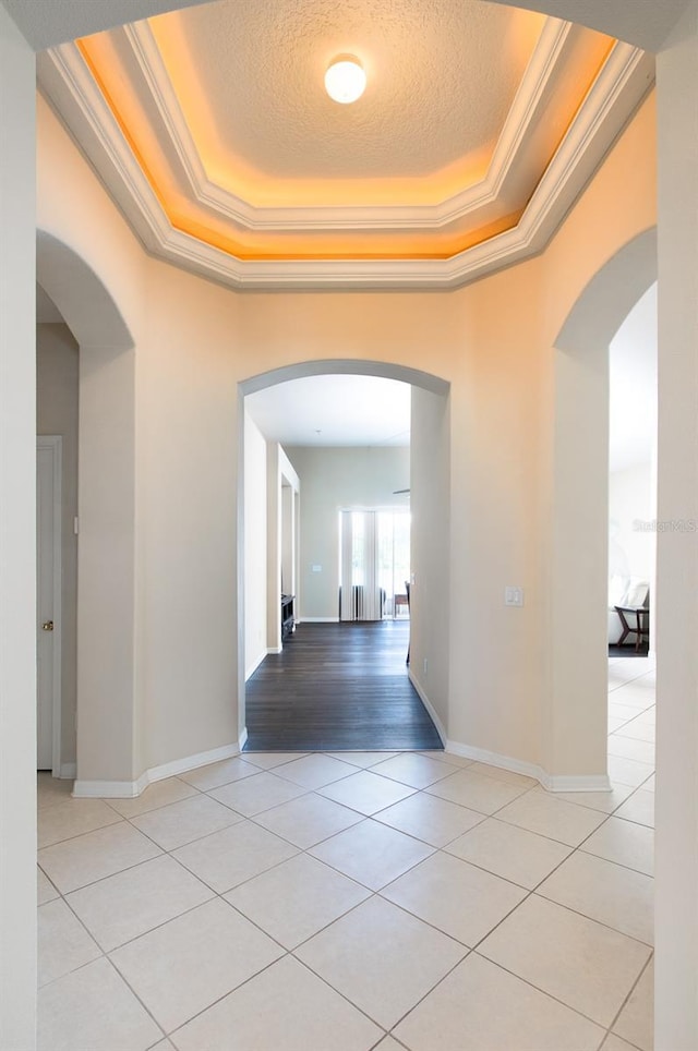 hall featuring light tile patterned floors, ornamental molding, a tray ceiling, arched walkways, and baseboards
