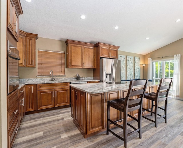 kitchen with light stone countertops, a sink, stainless steel appliances, brown cabinetry, and a center island