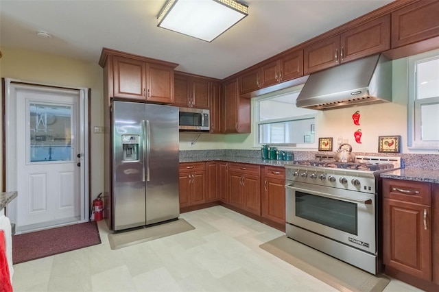 kitchen featuring brown cabinets, dark stone counters, stainless steel appliances, light floors, and under cabinet range hood