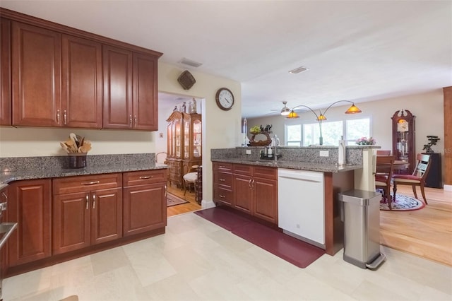 kitchen featuring a peninsula, white dishwasher, light floors, a sink, and dark stone countertops