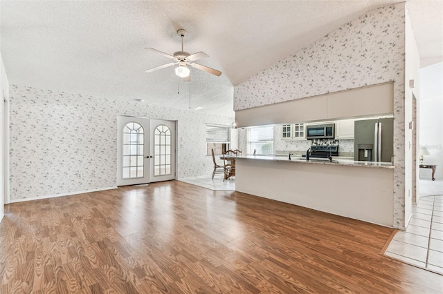 unfurnished living room with high vaulted ceiling, a ceiling fan, a textured ceiling, wallpapered walls, and wood finished floors