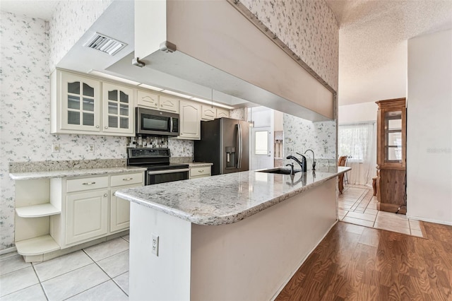 kitchen with a sink, open shelves, a peninsula, wallpapered walls, and appliances with stainless steel finishes