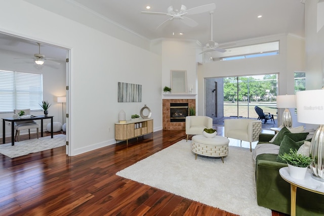 living room with a tiled fireplace, ornamental molding, a ceiling fan, a high ceiling, and dark wood-type flooring