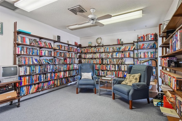 sitting room featuring ceiling fan, bookshelves, carpet flooring, visible vents, and a textured ceiling