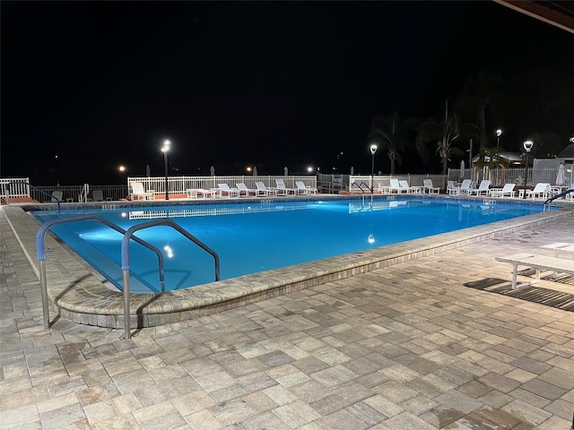 pool at night featuring a patio area, fence, and a community pool