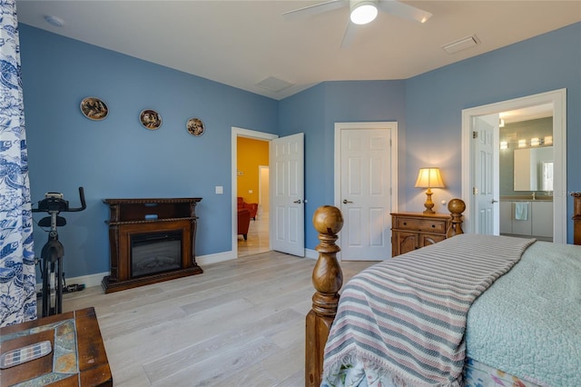 bedroom featuring ensuite bath, a glass covered fireplace, a ceiling fan, visible vents, and wood finished floors
