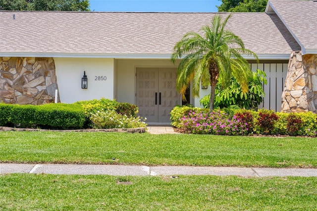 entrance to property with a lawn, roof with shingles, stucco siding, and stone siding