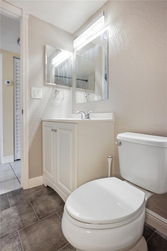 half bathroom featuring vanity, toilet, a textured wall, baseboards, and tile patterned flooring