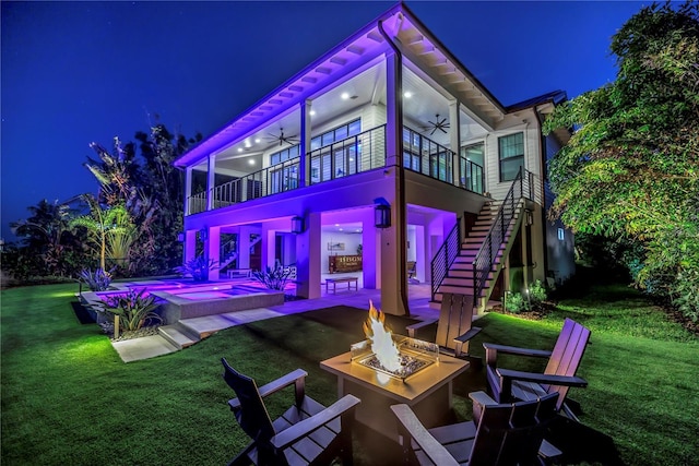 back of house at night featuring stairs, an outdoor fire pit, a ceiling fan, a patio, and a yard