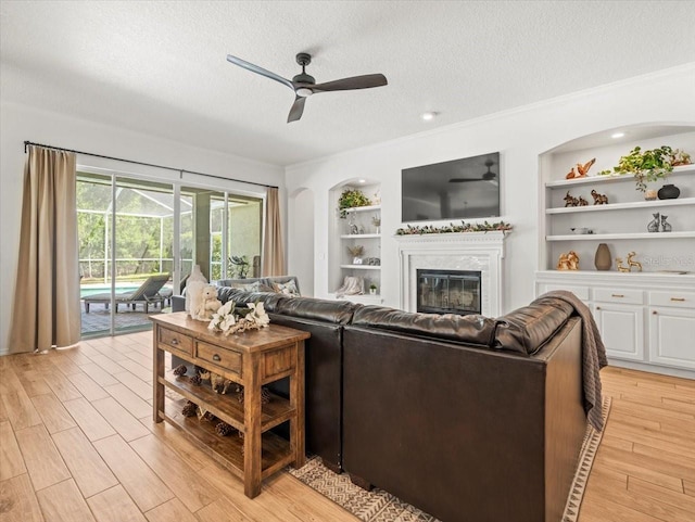 living area featuring a textured ceiling, ceiling fan, light wood-style floors, a glass covered fireplace, and built in shelves