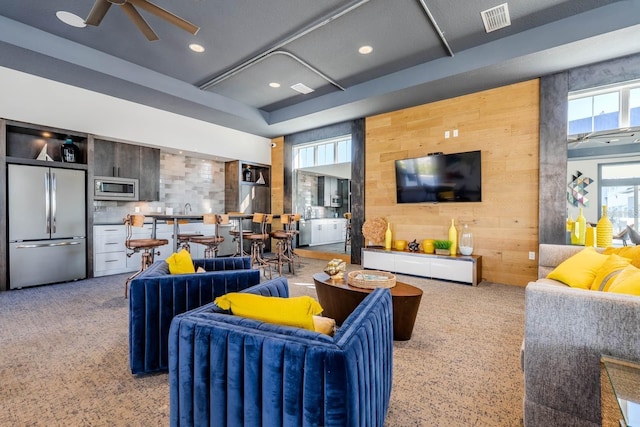 carpeted living room with ceiling fan, wood walls, and visible vents