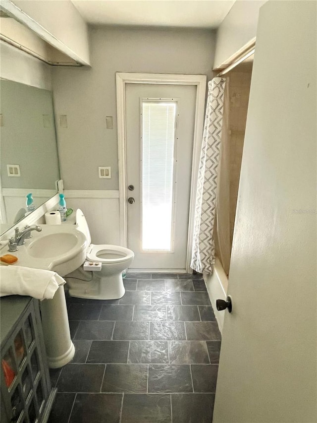 full bath with toilet, a wainscoted wall, a sink, and stone finish flooring