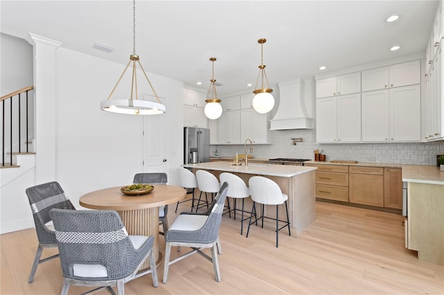 kitchen featuring custom exhaust hood, a kitchen island with sink, light wood-style flooring, backsplash, and a sink