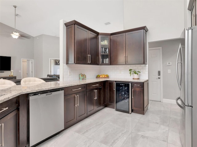 kitchen featuring appliances with stainless steel finishes, beverage cooler, dark brown cabinetry, light stone countertops, and backsplash