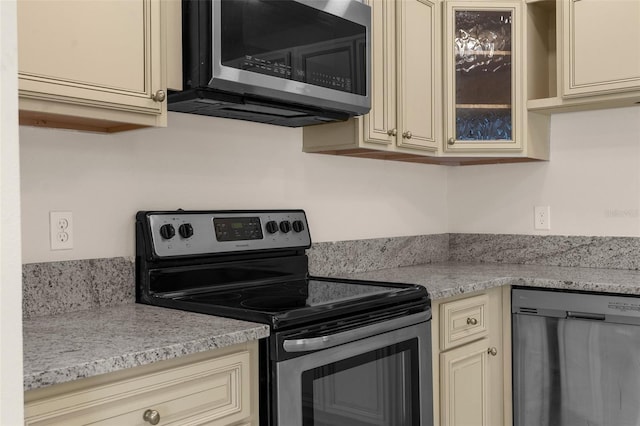 kitchen with appliances with stainless steel finishes, glass insert cabinets, and cream cabinets