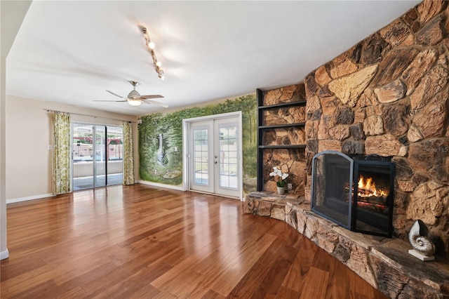 unfurnished living room featuring a stone fireplace, french doors, a ceiling fan, baseboards, and wood finished floors