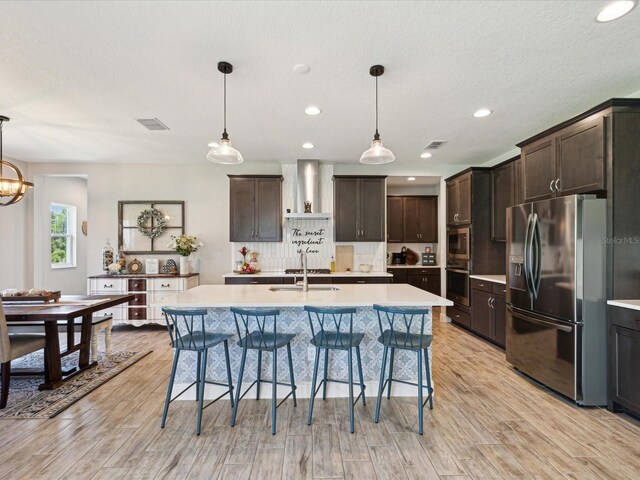 kitchen with dark brown cabinetry, light countertops, wall chimney exhaust hood, stainless steel appliances, and a kitchen bar