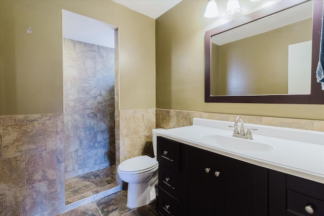 bathroom with a tile shower, vanity, toilet, a wainscoted wall, and tile walls
