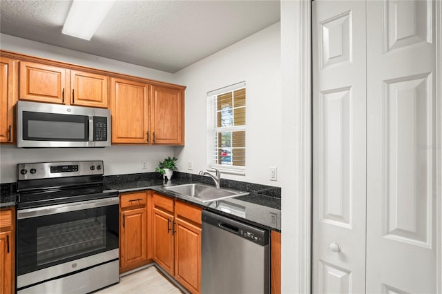kitchen featuring brown cabinetry, a textured ceiling, a sink, dark stone countertops, and stainless steel appliances