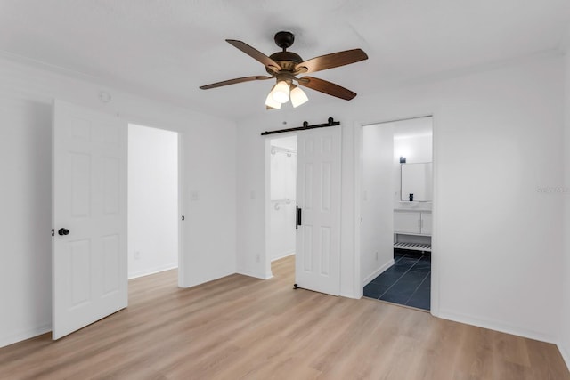 unfurnished bedroom featuring a barn door, wood finished floors, a spacious closet, a ceiling fan, and ensuite bath