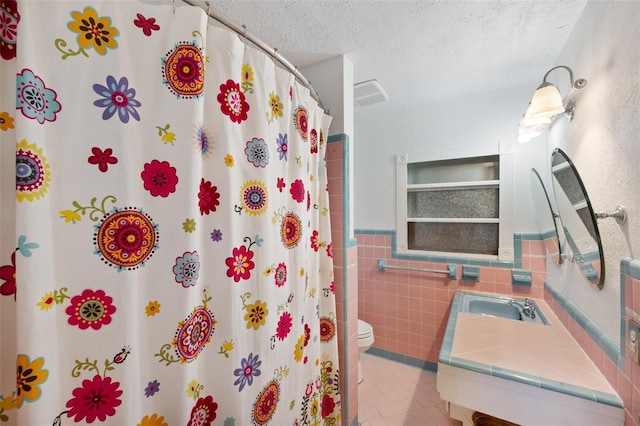 full bathroom with wainscoting, tile patterned flooring, toilet, a textured ceiling, and tile walls