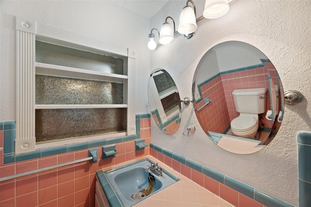 bathroom featuring a textured wall, wainscoting, a sink, toilet, and tile walls