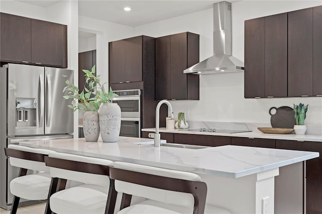 kitchen featuring wall chimney range hood, appliances with stainless steel finishes, dark brown cabinetry, and a kitchen bar