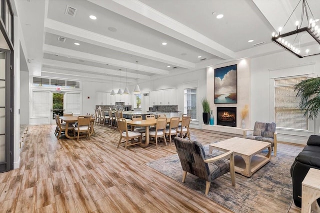 living room with a warm lit fireplace, recessed lighting, light wood-type flooring, an inviting chandelier, and beam ceiling