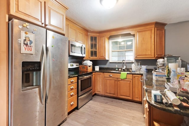 kitchen featuring glass insert cabinets, dark stone counters, stainless steel appliances, a sink, and light wood-type flooring