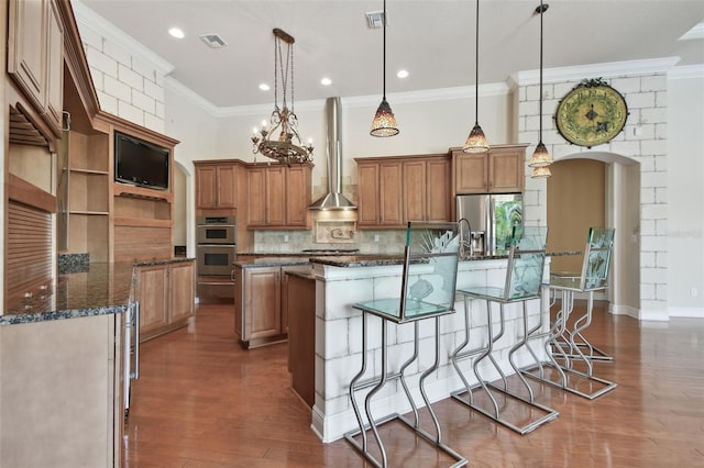 kitchen featuring visible vents, ornamental molding, an island with sink, wall chimney range hood, and appliances with stainless steel finishes