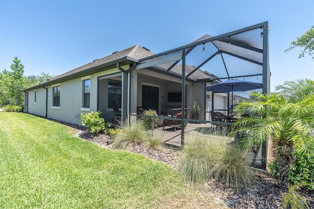 rear view of property featuring roof with shingles, a patio area, glass enclosure, a yard, and stucco siding