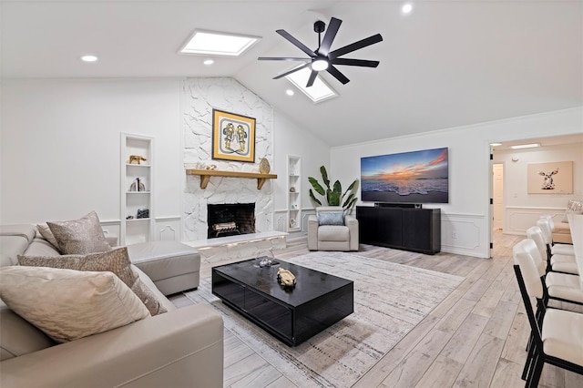 living area featuring vaulted ceiling with skylight, a ceiling fan, a fireplace, light wood-type flooring, and built in shelves