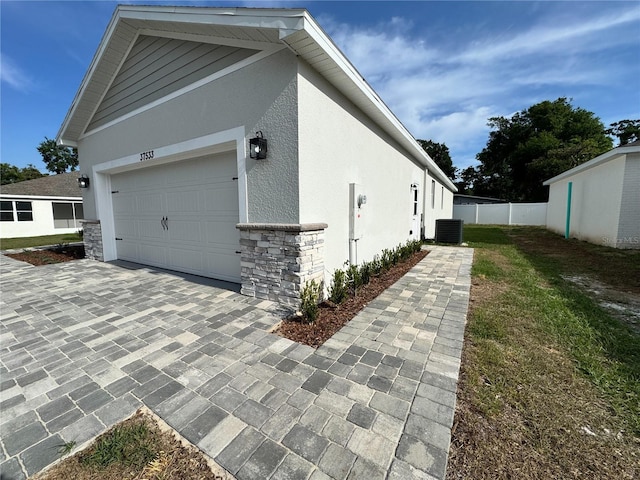 view of side of home featuring stucco siding, fence, central AC unit, driveway, and stone siding