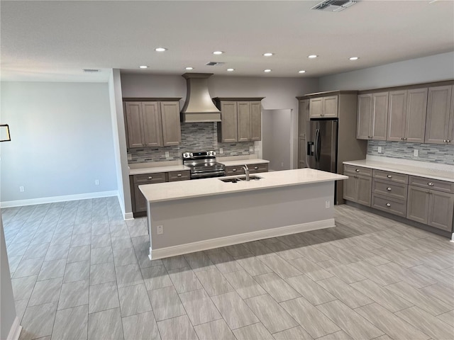 kitchen featuring tasteful backsplash, custom range hood, a sink, stainless steel appliances, and a center island with sink