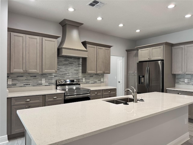 kitchen with decorative backsplash, a sink, appliances with stainless steel finishes, premium range hood, and a center island with sink