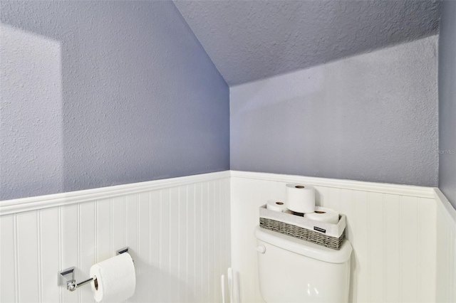 bathroom featuring a textured wall, wainscoting, toilet, a textured ceiling, and vaulted ceiling