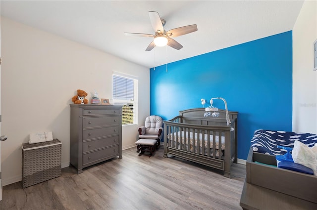 bedroom with baseboards, a ceiling fan, a nursery area, and wood finished floors