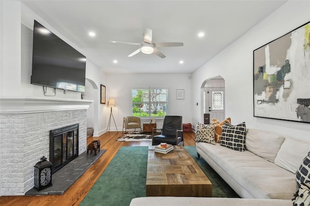 living area with ceiling fan, arched walkways, a brick fireplace, wood finished floors, and baseboards