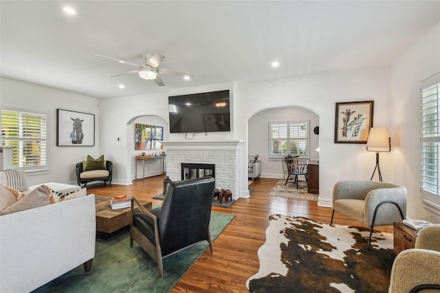 living room with ceiling fan, a brick fireplace, arched walkways, recessed lighting, and wood finished floors