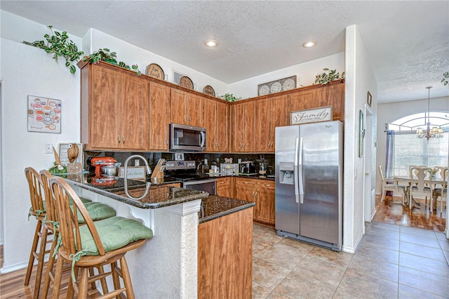 kitchen with a textured ceiling, appliances with stainless steel finishes, brown cabinets, dark stone countertops, and a peninsula