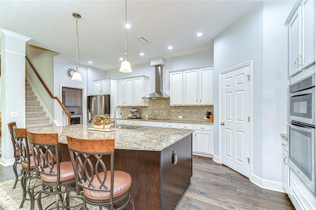kitchen with wall chimney range hood, visible vents, appliances with stainless steel finishes, dark wood finished floors, and a sink
