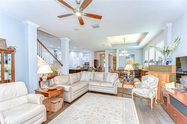 living room with a raised ceiling, ceiling fan with notable chandelier, decorative columns, wood finished floors, and visible vents