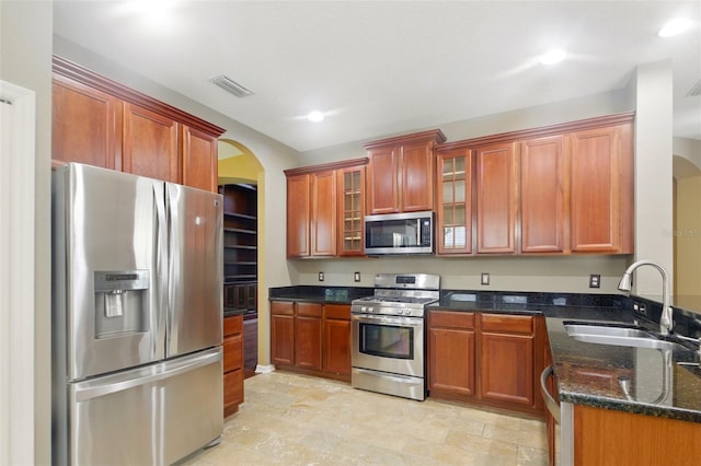 kitchen with a sink, glass insert cabinets, arched walkways, dark stone countertops, and stainless steel appliances