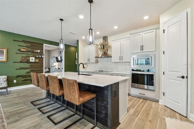 kitchen with wall chimney range hood, appliances with stainless steel finishes, a sink, light countertops, and light wood finished floors