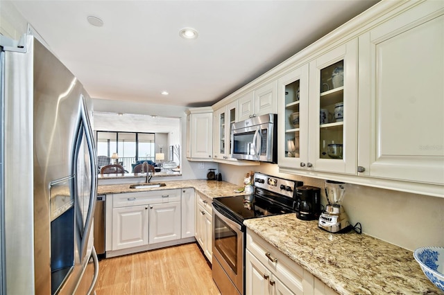kitchen featuring glass insert cabinets, a sink, stainless steel appliances, light wood-type flooring, and light stone countertops