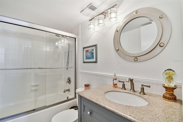 full bath featuring toilet, visible vents, vanity, a wainscoted wall, and combined bath / shower with glass door