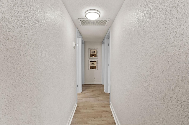 hallway featuring baseboards, a textured wall, visible vents, and light wood-style flooring