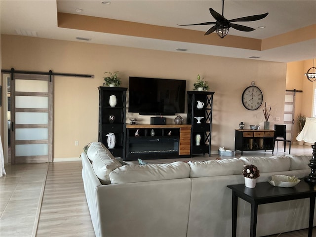 living room featuring ceiling fan, a barn door, light wood finished floors, baseboards, and a tray ceiling